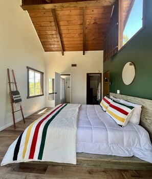 Ein Schlafzimmer mit Holzbalkendecke, einem Bett mit weißer Bettwäsche, einer gestreiften Decke und Blick ins Freie.