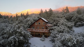 Una cabaña de madera con balcón, rodeada de árboles nevados y montañas al fondo.