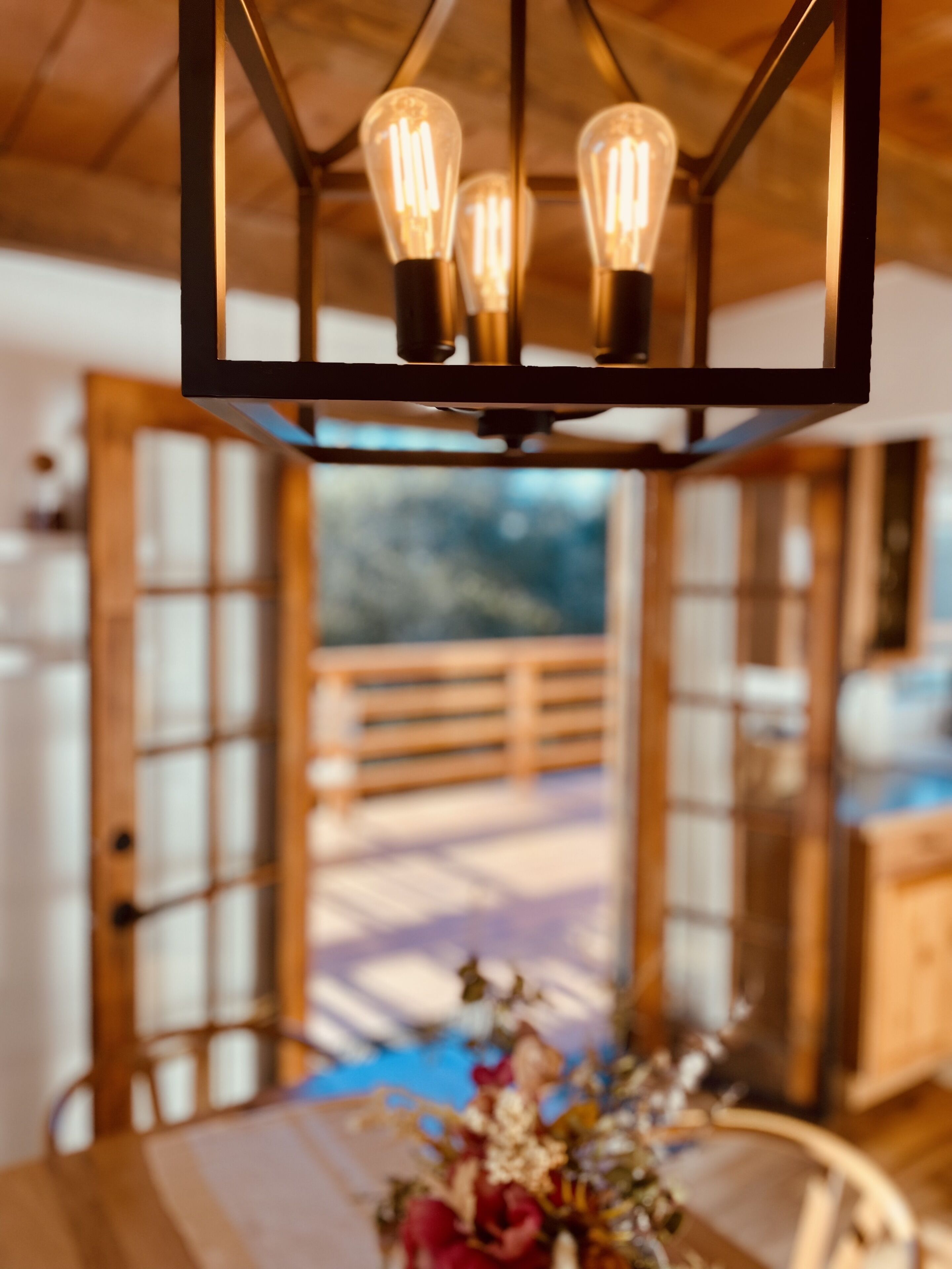 A dining area with a wooden table and chairs, a chandelier with three light bulbs, and a view of a wooden deck through a glass door.