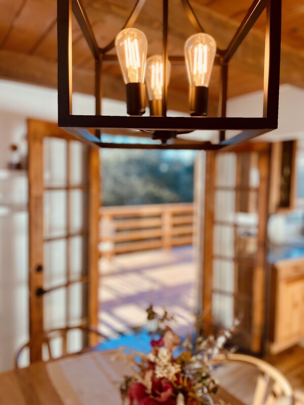 Un coin repas avec une table et des chaises en bois, un lustre à trois ampoules et une vue sur une terrasse en bois, visible à travers une porte vitrée.