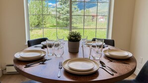 Une table à manger dressée pour quatre personnes, avec assiettes, couverts et verres, placée devant une fenêtre offrant une vue sur la verdure et les montagnes.