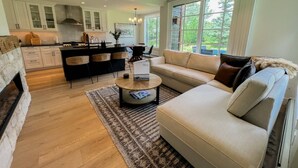 Un salon moderne avec un canapé beige, une table basse et un tapis. On y trouve également un coin cuisine avec des meubles blancs et un pare-feu en pierre.