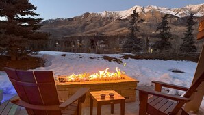 Une terrasse en bois avec un foyer en pierre et deux fauteuils Adirondack, le tout donnant sur un paysage montagneux enneigé.