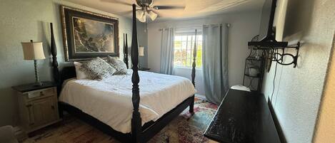 A bedroom with a four-poster bed, a ceiling fan, a painting on the wall, and a desk with a lamp.