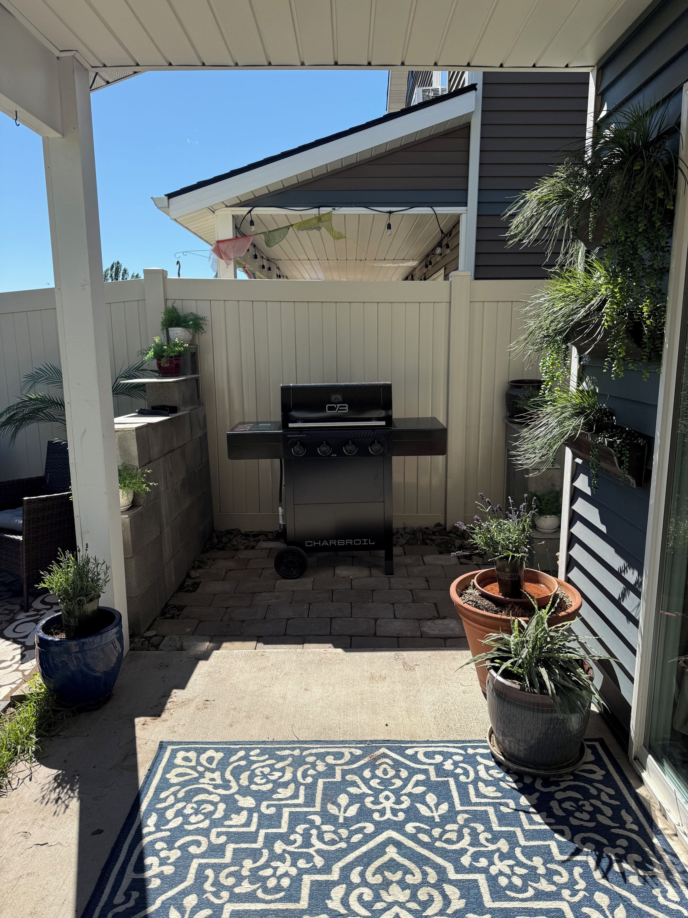 A covered patio area with a barbecue grill, potted plants, and a patterned rug.