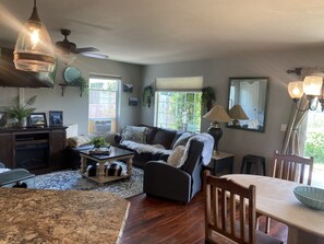 Un salon avec un canapé marron foncé, une table basse et une table à manger avec des chaises. On y trouve également un ventilateur de plafond, une lampe et une fenêtre avec des stores.