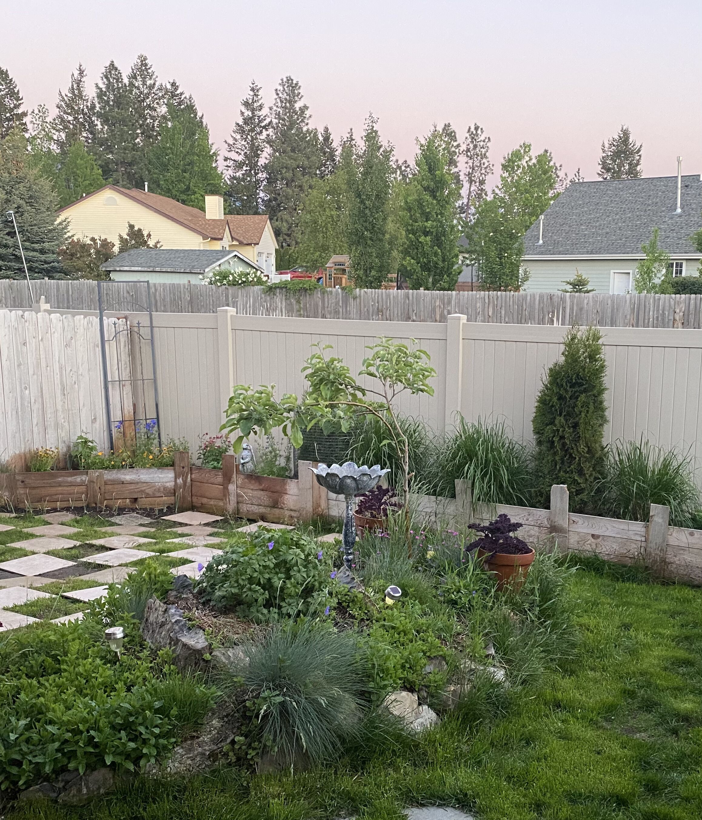 Ein Garten mit Holzzaun, verschiedenen Pflanzen und Blumen sowie einem gepflasterten Weg.