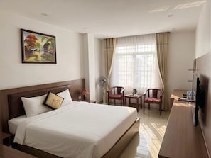 Quarto de hotel com uma cama grande, uma escrivaninha com cadeiras, uma televisão e uma janela com cortinas.