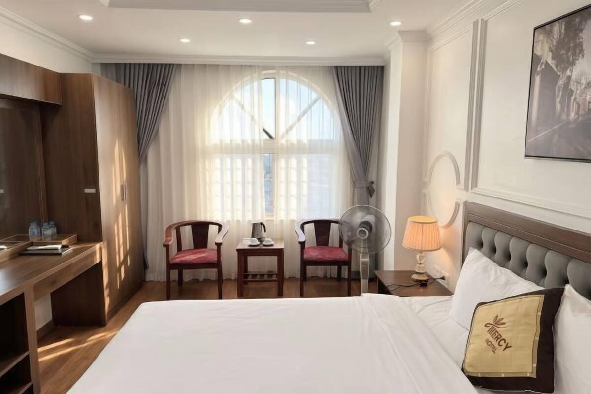 Habitació d'hotel amb un llit, dues cadires, un escriptori, un ventilador, una làmpada i una finestra amb cortines.