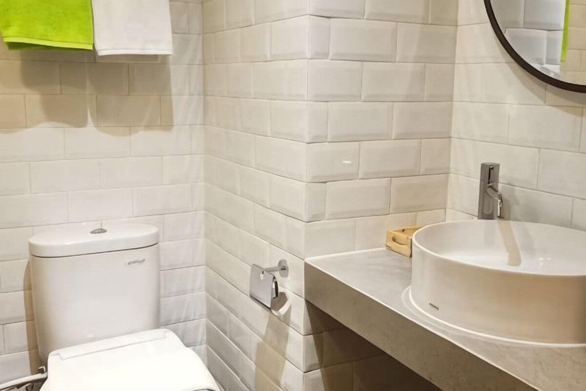 Un bagno con pareti piastrellate di bianco, un water, un lavandino con uno specchio e un portasciugamani con un asciugamano verde.