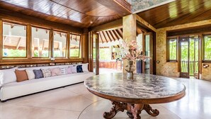 A spacious room with a round marble table, a white sofa with patterned cushions, and large windows offering a view of greenery outside. - AV Villa Canggu Hearts (Canggu)