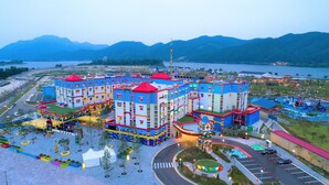 Un complejo hotelero colorido con varias edificaciones, un estacionamiento y vista a un lago y montañas al fondo.