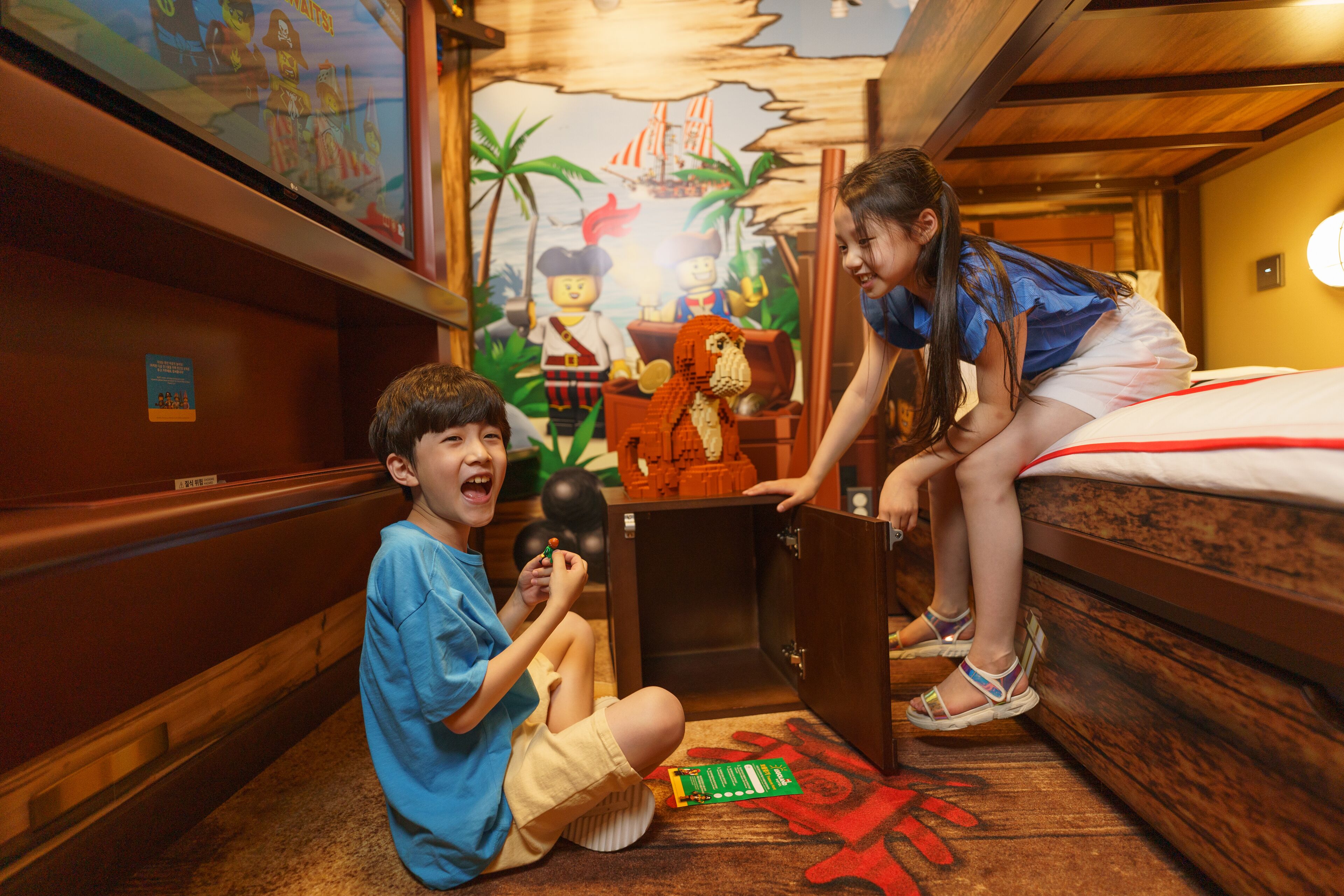A room with wooden walls and a mural depicting pirates. There is a bed with a red and white bedspread, and a small table with a green object on it.