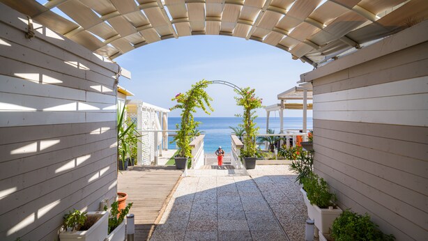 A covered outdoor area with a view of the sea, wooden flooring, and potted plants.