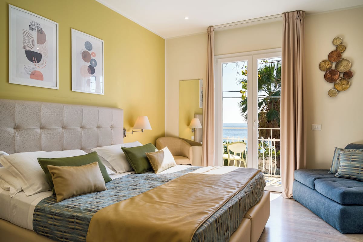 Habitación de hotel con una cama grande, un sofá, un balcón con vistas al mar y obras de arte en las paredes.