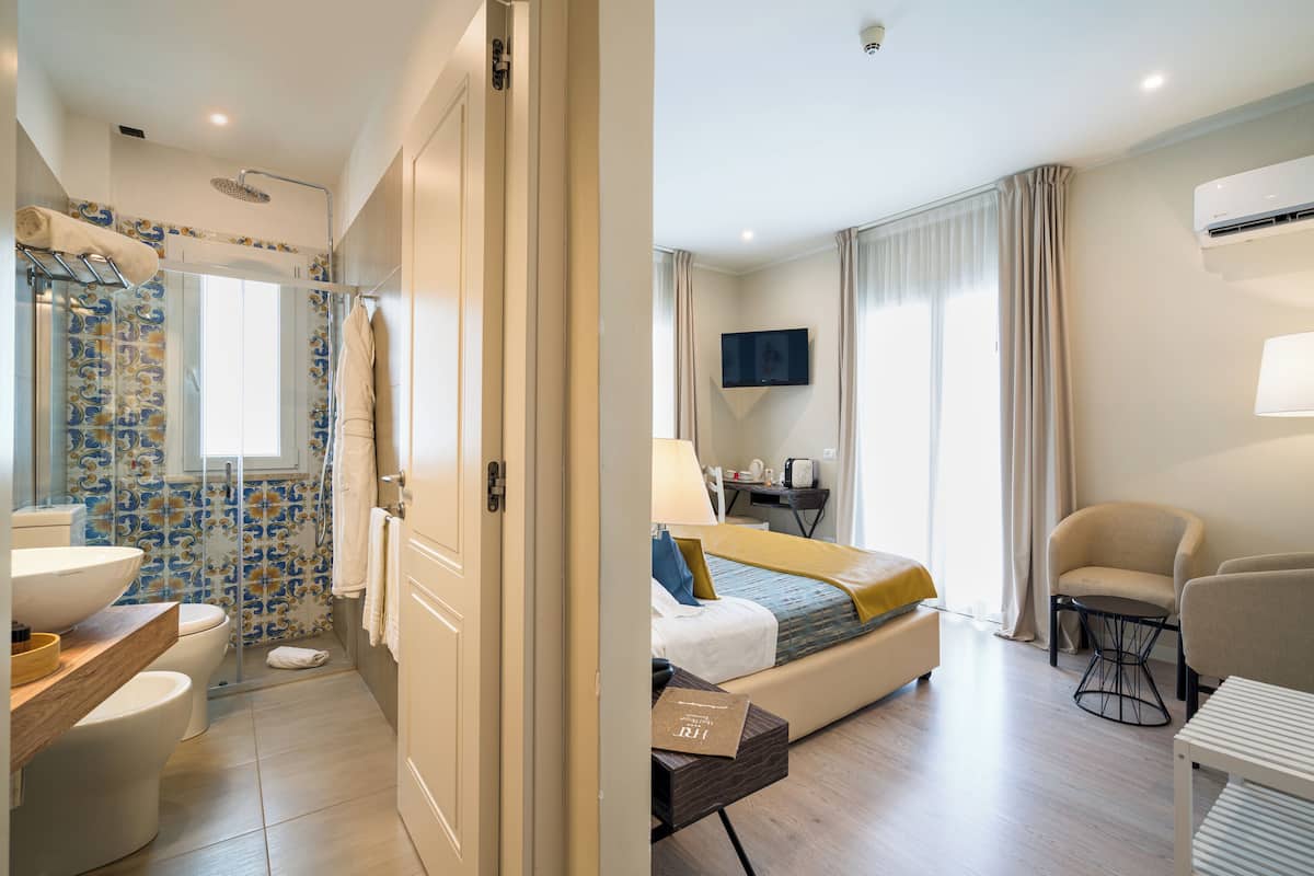 Habitación de hotel moderna con ducha, inodoro y lavamanos; una cama con televisor y escritorio; y una silla.