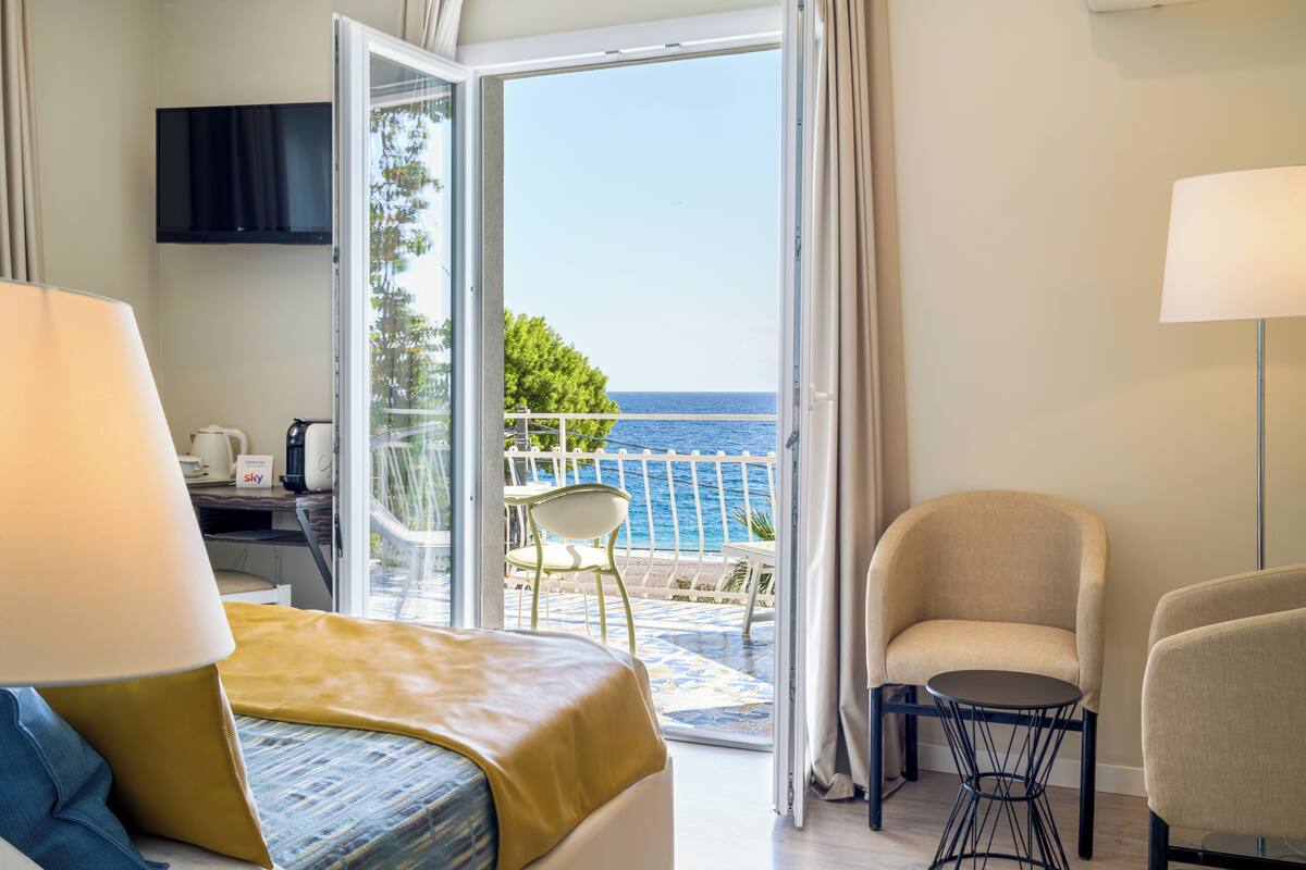 Habitación de hotel con cama, escritorio, silla, mesa y televisor. La habitación tiene un balcón con vista al mar.