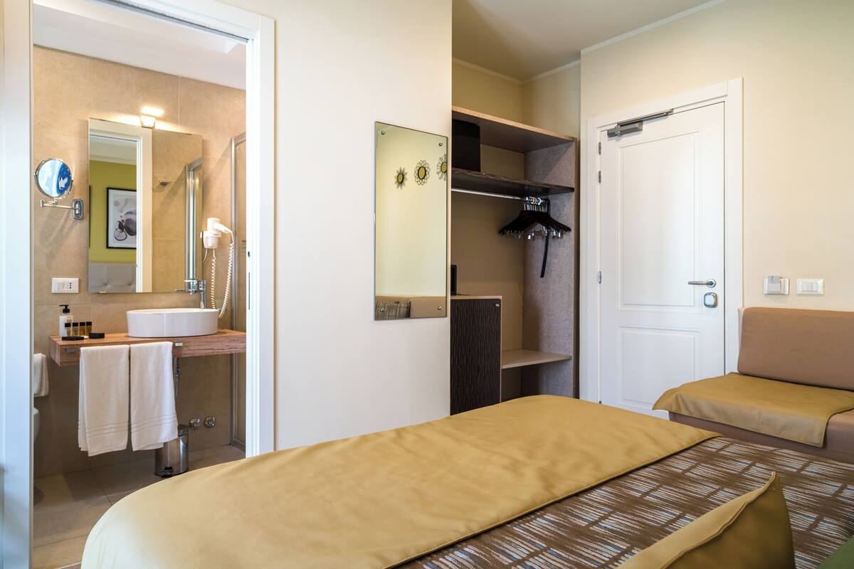 Habitación de hotel con cama, mesita de noche, espejo, baño con lavamanos y ducha.