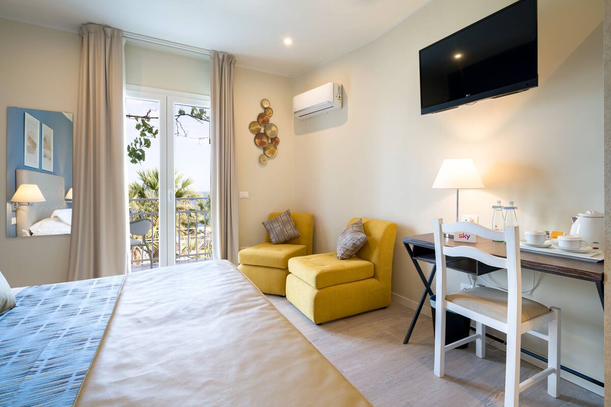Habitación de hotel con una cama, dos sillones amarillos, un escritorio blanco con una silla, un televisor de pantalla plana y un equipo de aire acondicionado.