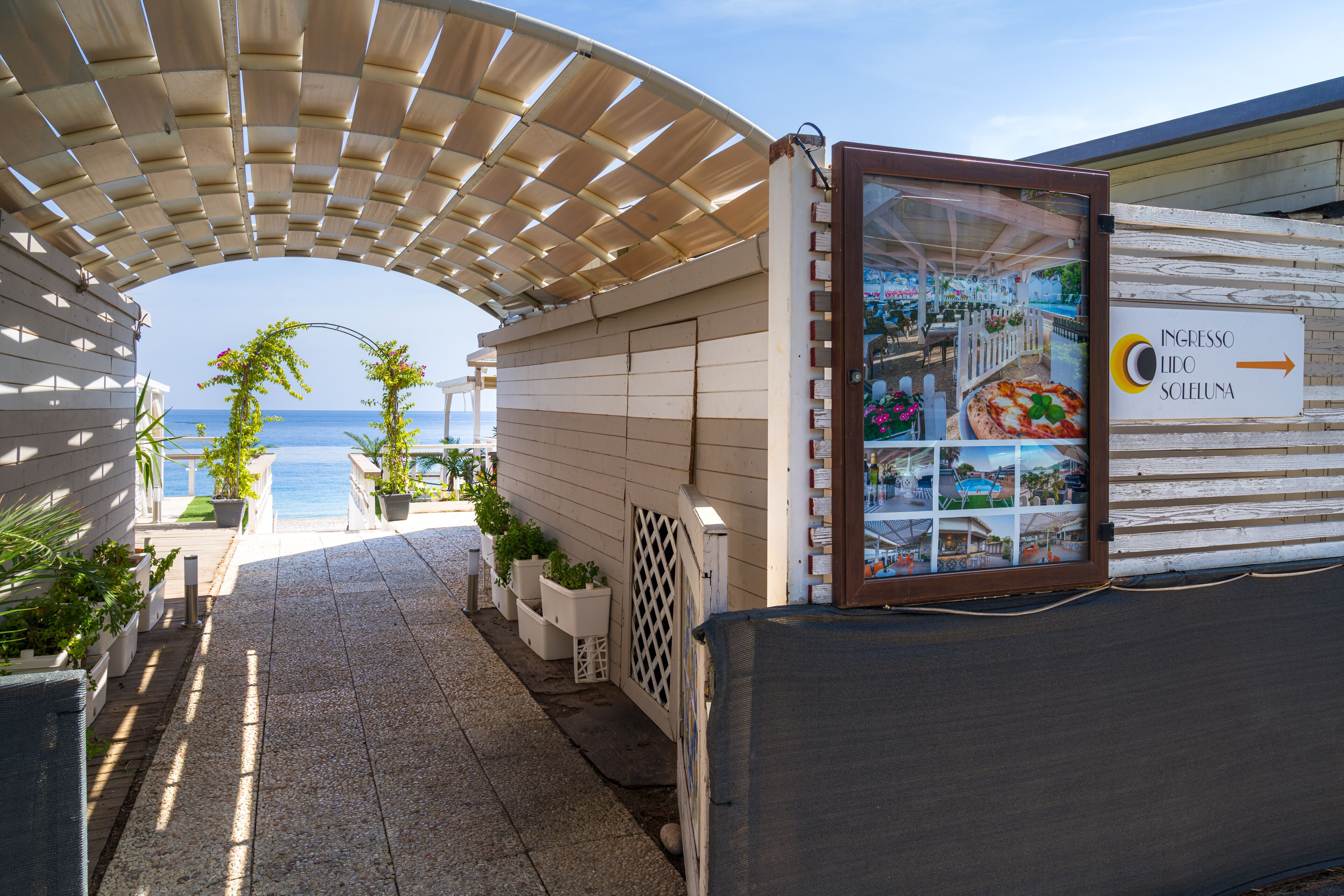 A pathway leading to a beachside area with a sign indicating 'Ingresso Lido Scelena' and a view of the sea.