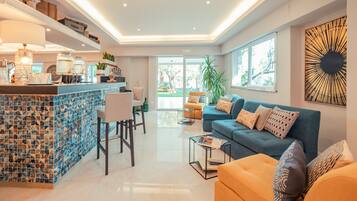 Ein modernes Wohnzimmer mit einem blauen Sofa, einem orangen Sofa und einem Couchtisch. Es gibt eine Küchenzeile mit einer Bar und einem Esstisch.