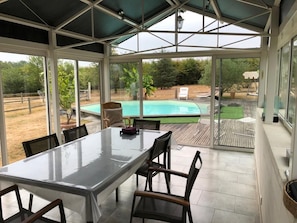 En täckt uteplats med ett matbord och stolar, med utsikt över en swimmingpool och en trädgård.