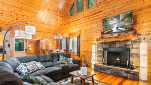 L’intérieur chaleureux d’un chalet avec une cheminée, un téléviseur fixé au-dessus et un coin repas à l’arrière-plan.