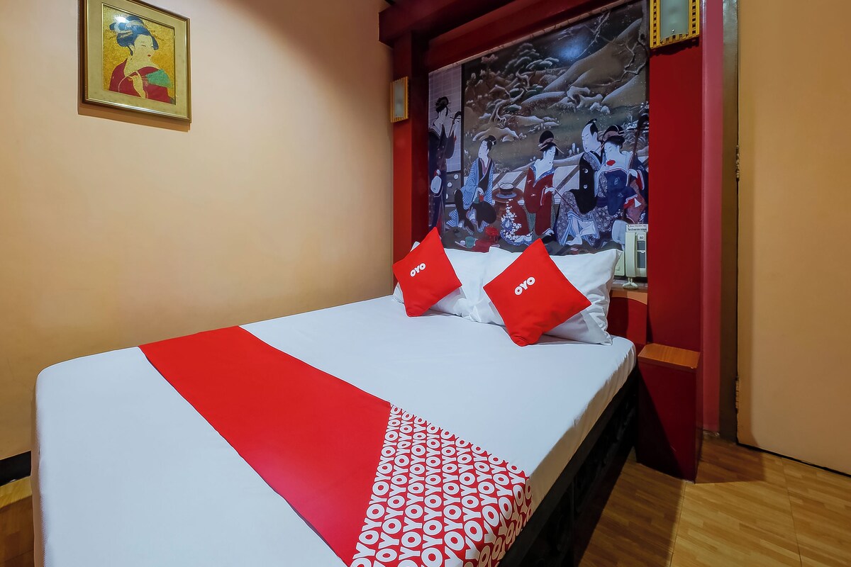 A hotel room with a bed, red and white bedding, a framed picture on the wall, and a large tapestry depicting a traditional scene.