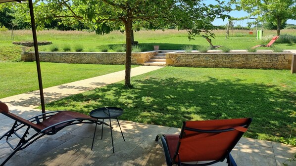A patio area with two chairs and a small table, a stone wall, and a grassy lawn with a playground in the background.