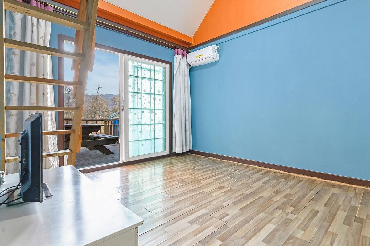 A room with a wooden floor, a TV on a stand, a large window with curtains, and an air conditioning unit on the wall.