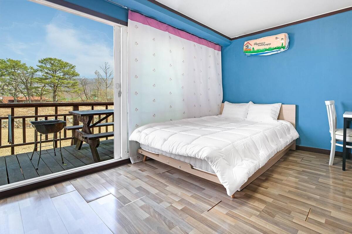 A bedroom with a bed, a desk, and a chair. There is a window with a view of trees and a building.