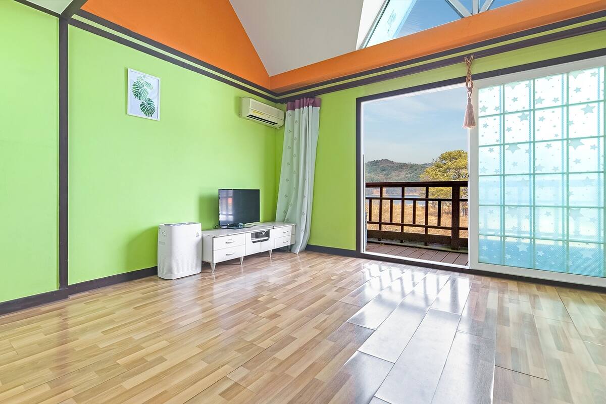 A room with a wooden floor, a TV on a white cabinet, an air conditioner, and a large window with a view of a landscape.