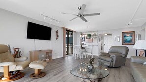 A modern living room with a flat-screen TV, a ceiling fan, a glass coffee table, and a dining area with white chairs.