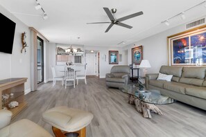 Un salon moderne avec un ventilateur de plafond, une table basse en verre, un canapé beige et un coin repas avec des chaises blanches.
