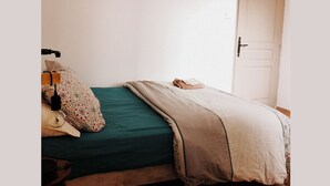 Un lit simple avec une couvre-lit verte et une couverture beige, une table de chevet avec une lampe, et une porte sur la droite.