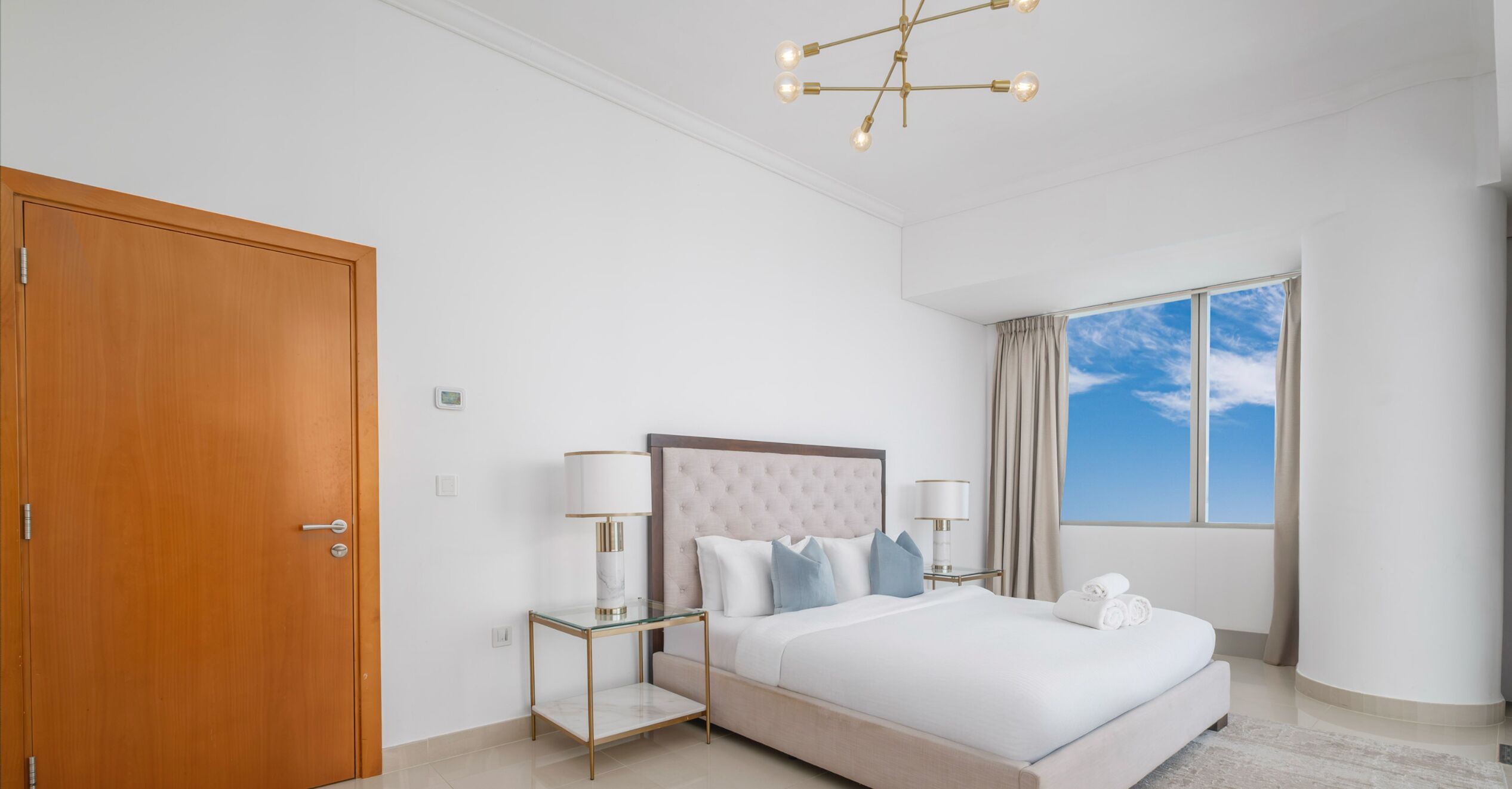 Habitación de hotel moderna con una cama grande, una puerta de madera y vista al cielo a través de una ventana.