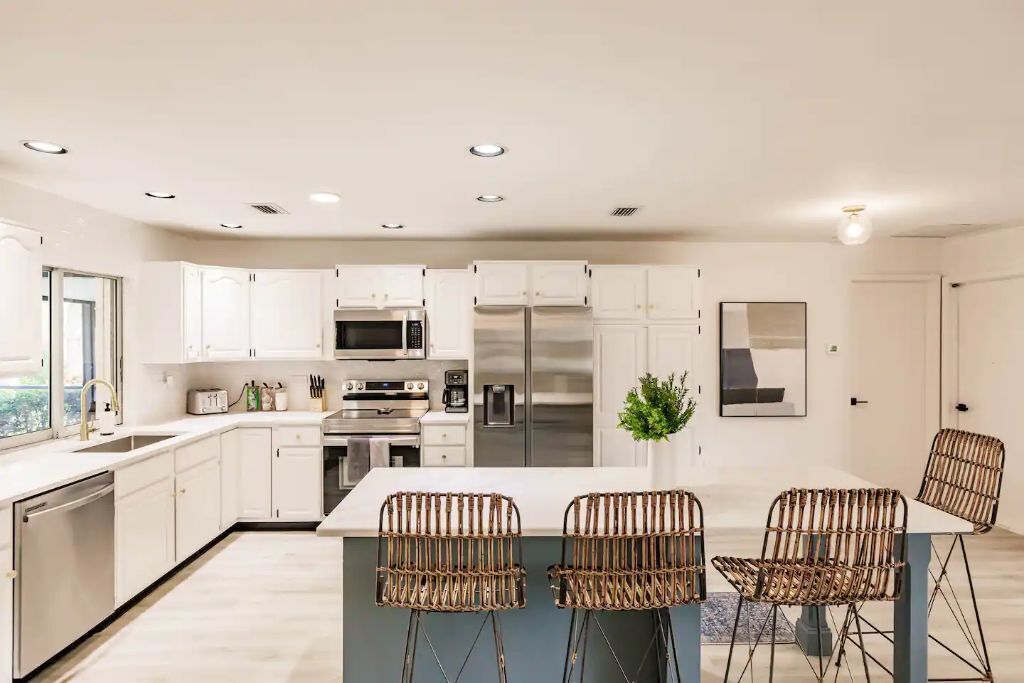 A modern kitchen with white cabinetry, stainless steel appliances, and a central island with wicker chairs.