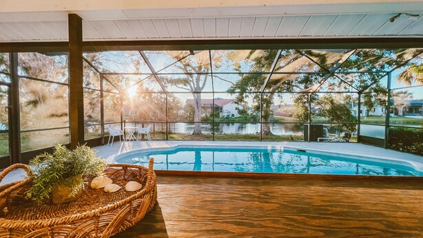 Zona de piscina con vista a un lago y árboles, una cesta de mimbre con conchas y una mesa de madera.