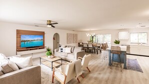 Una sala de estar moderna con televisor, sofá y mesa de centro. Hay una zona de comedor con sillas y una mesa. El piso es de madera y las paredes son blancas.