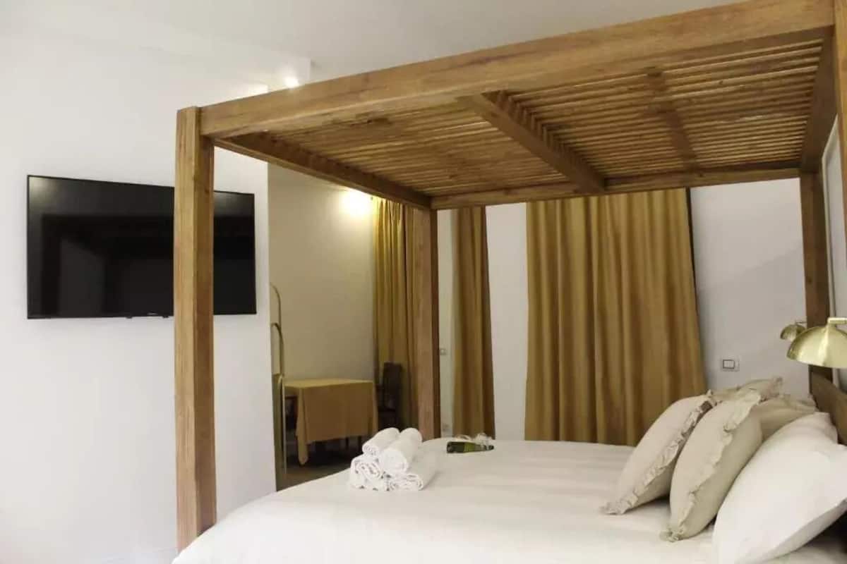A bedroom with a wooden ceiling structure, a bed with white linens, a flat-screen TV mounted on the wall, and a window with curtains.