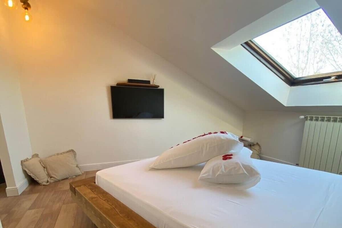 A bedroom with a skylight, a wooden platform bed, white bedding, and a mounted TV.