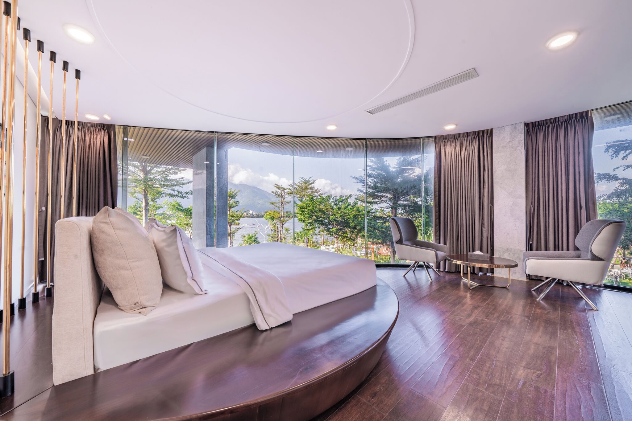 A modern bedroom with a curved bed, a small round table, two chairs, and large windows offering a view of trees and mountains.