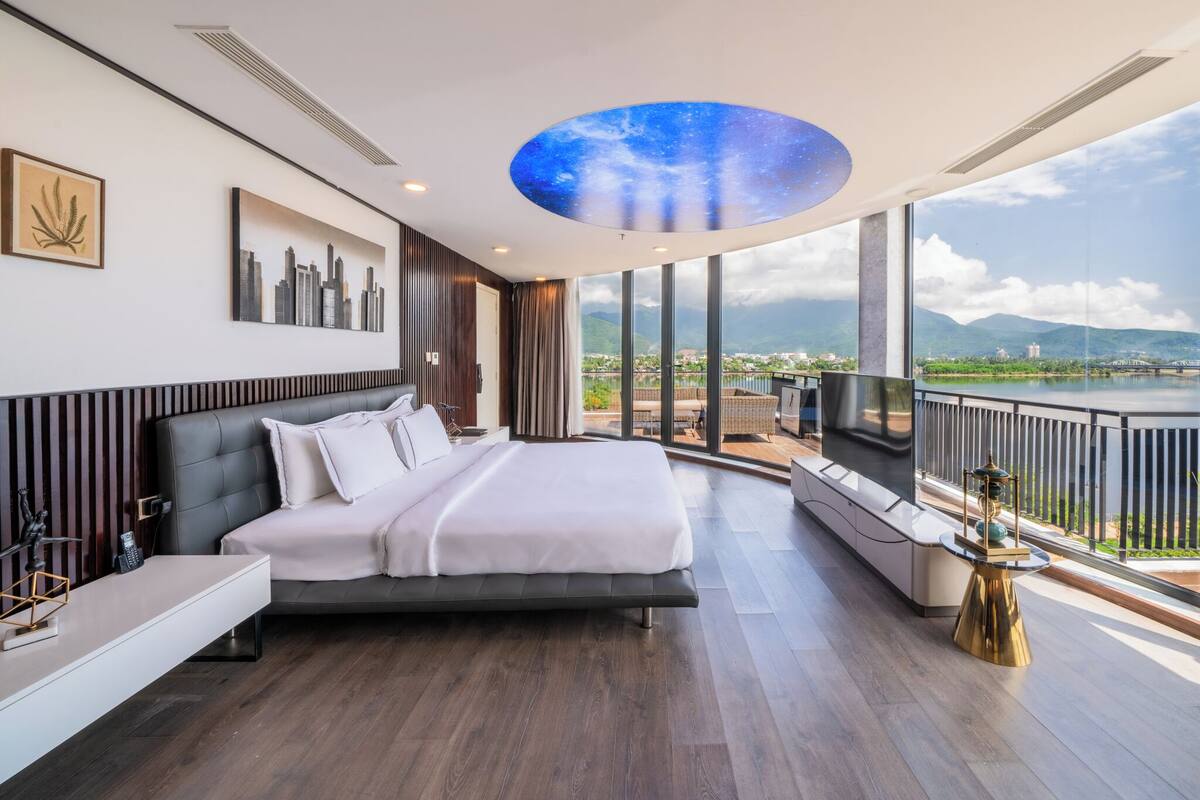A modern bedroom with a large bed, a cityscape painting, a ceiling with a starry sky design, and a view of a lake and mountains.