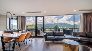 A spacious living area with a large window offering a mountain view, a dining area with a glass table and chairs, and a modern black leather sofa. - Cami Riverside Resort Đà Nẵng (Da Nang)