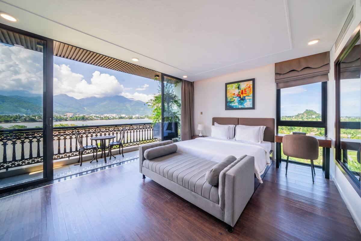 A spacious bedroom with a large bed, a sofa, and a desk. It features a balcony with a view of a mountain and a river.