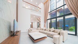A spacious living room with a large sectional sofa, a flat-screen TV on a wooden console, and a large window with curtains. - Cami Riverside Resort Đà Nẵng (Da Nang)
