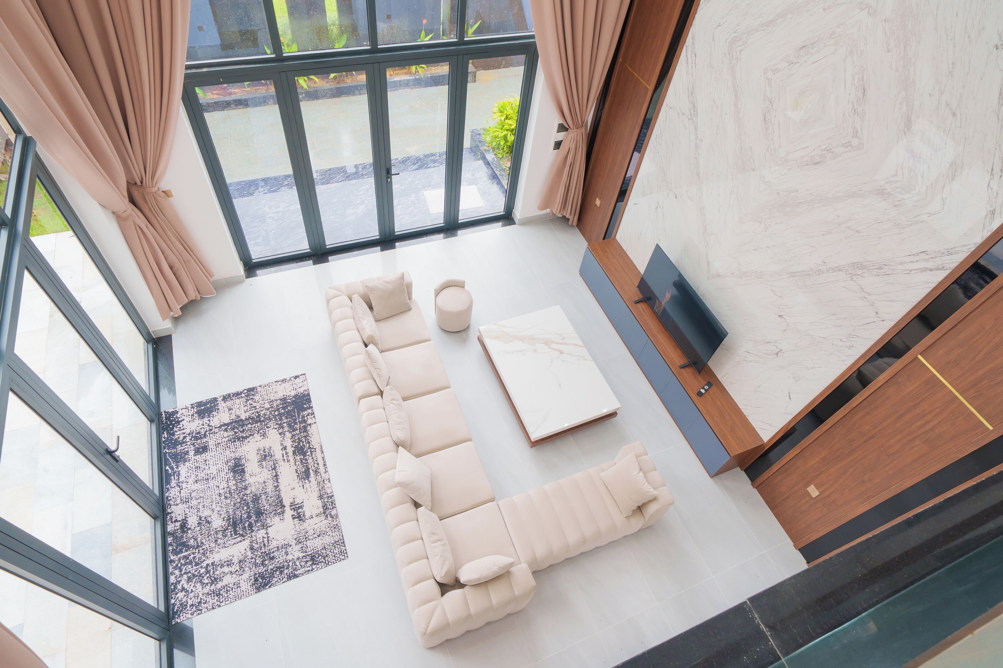 A modern living room with a sectional sofa, a coffee table, and a TV. There is a large window with curtains, a patterned rug, and a marble wall.
