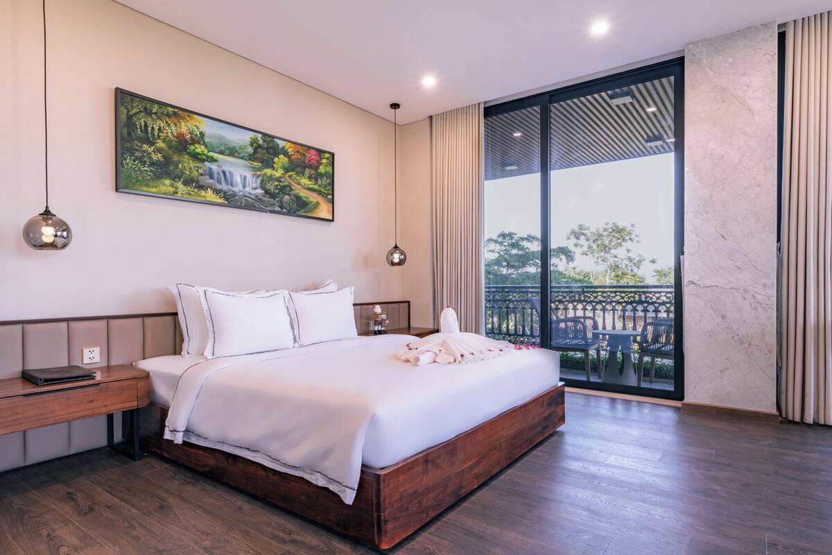 A modern bedroom with a large bed, a nightstand, a painting, and a balcony with outdoor furniture.
