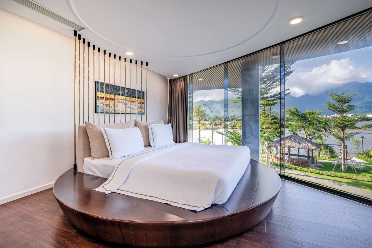 A modern bedroom with a curved bed, a large window with a view of greenery and mountains, and a painting on the wall.