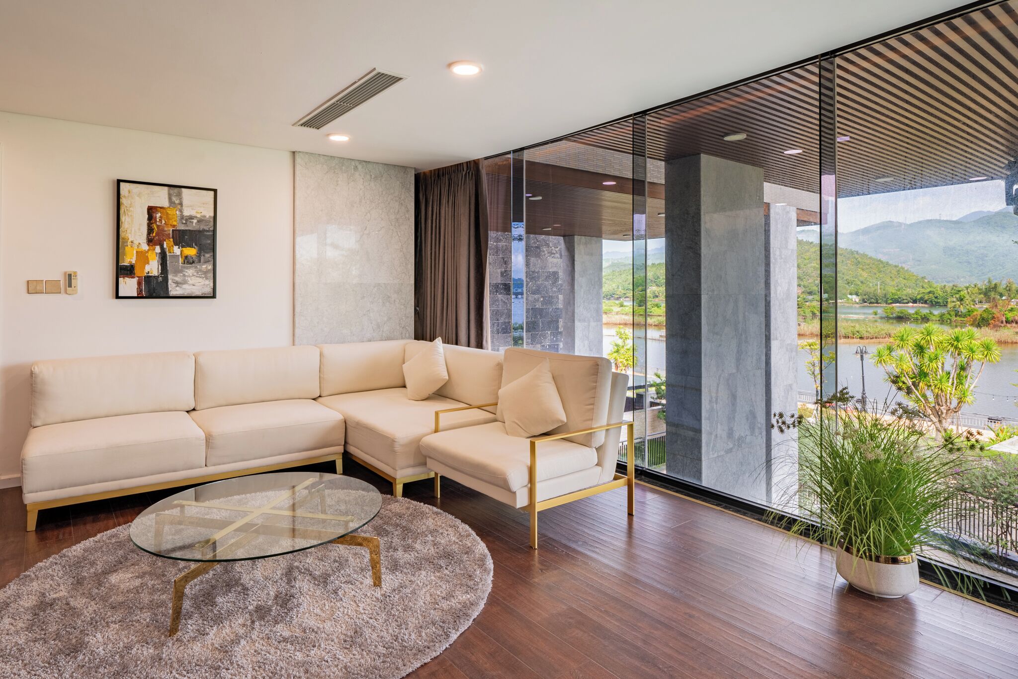 Uma sala de estar moderna com um sofá seccional, uma mesa de centro de vidro e vista para um lago e montanhas através de grandes janelas.
